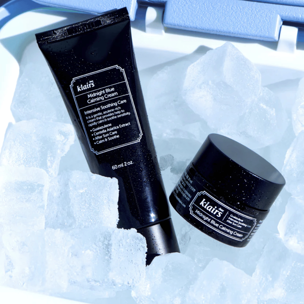 Eine Tube und ein Glas von Klairs Midnight Blue Calming Cream liegen auf Eiswürfeln, mit Fokus auf beruhigende und feuchtigkeitsspendende Eigenschaften, vor einem blauen Hintergrund.