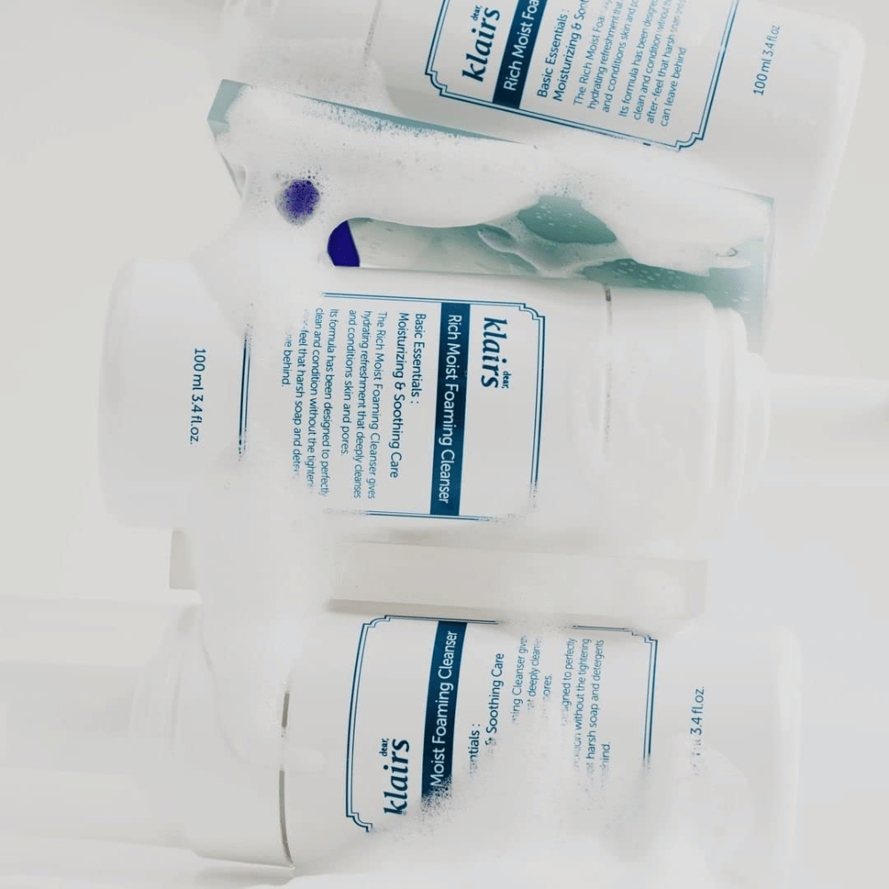 Drei Flaschen von Klairs Rich Moist Foaming Cleanser bedeckt mit Schaum und Wassertropfen, auf einer weißen Fläche platziert.