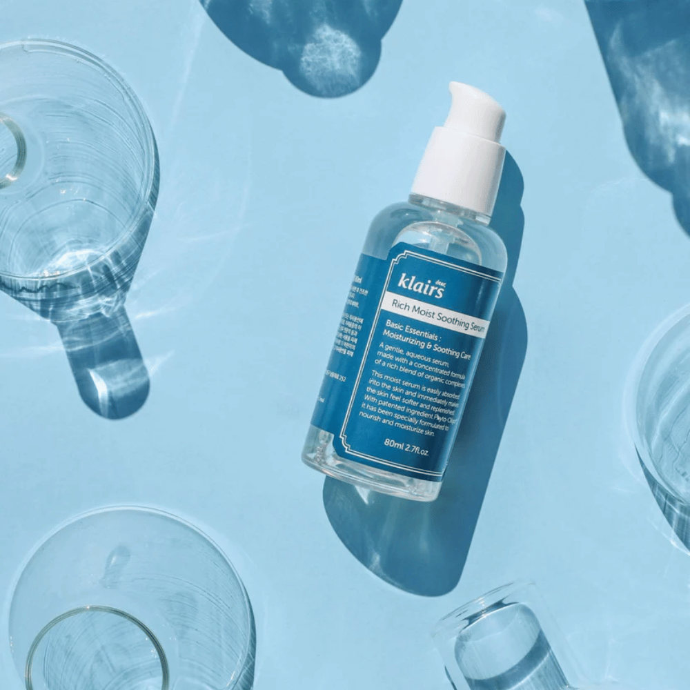 Eine Flasche Klairs Rich Moist Soothing Serum zwischen Wassergläsern mit Reflexionen auf einem blauen Hintergrund.