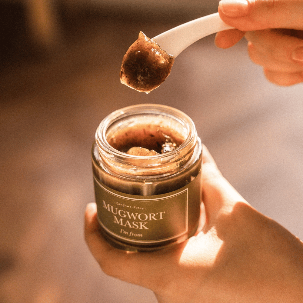 Nahaufnahme einer Hand, die I'm From Mugwort Mask hält, während ein Löffel die kräuterbasierte Gelmaske aus dem Behälter hebt, im warmen natürlichen Licht.