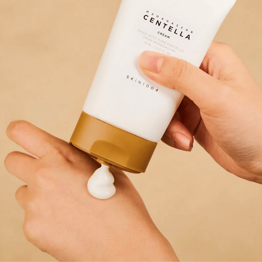 Das Bild zeigt eine Person, die eine Tube "Madagascar Centella Cream" von SKIN1004 hält. Es ist eine Hautcreme, die aus reiner Centella aus Madagaskar hergestellt wird, in der Größe 75 ml / 2.53 fl.oz. Die Creme wird auf die Hand aufgetragen und soll der Haut Nährstoffe und Feuchtigkeit spenden.