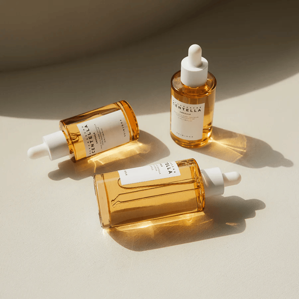 Das Bild zeigt drei Flaschen von "Madagascar Centella Asiatica 100 Ampoule". Sie sind auf einer Fläche platziert, auf der das Sonnenlicht eine schöne Reflexion durch den goldenen Inhalt in den Flaschen erzeugt. Das Produkt scheint Teil einer Hautpflegeserie zu sein, die Centella Asiatica verwendet, eine Zutat, die für ihre beruhigenden Eigenschaften bekannt ist. Das Design der Flaschen ist einfach und modern, was darauf hindeutet, dass es sich um eine Premium-Hautpflegeserie handelt.