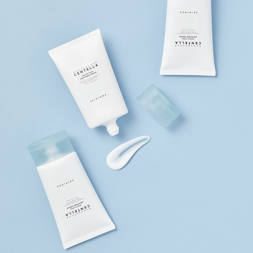 Zwei weiße Tuben mit SKIN1004 Centella Hautpflegeprodukten, ein Klecks Produkt und ein klarer blauer Verschluss, auf einem hellblauen Hintergrund.