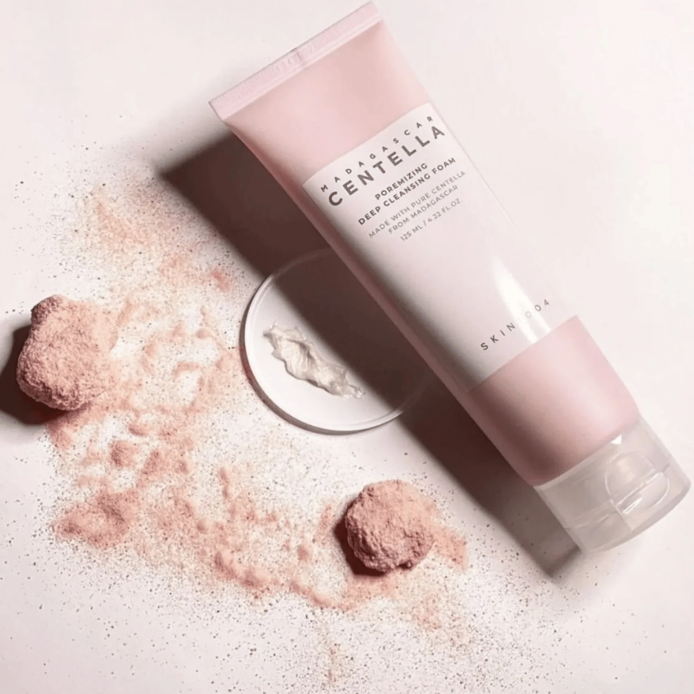Madagascar Centella Poremizing Deep Cleansing Foam wird in einer rosa Tube präsentiert. Die Tube liegt auf einem hellen Untergrund zusammen mit rosa Pulver, das ein Gefühl von Reinheit und Frische vermittelt. Das Produkt ist dafür ausgelegt, die Haut mit Hilfe von reiner Centella aus Madagaskar tief zu reinigen.