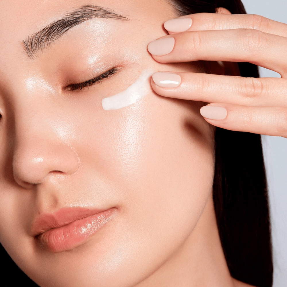 Eine Frau trägt vorsichtig eine weiße Augencreme unter dem Auge auf. Ihre Haut sieht strahlend und feucht aus, was die Wirkung der Augencreme betont. Dies unterstreicht das Produkt als sowohl feuchtigkeitsspendend als auch geeignet für die empfindliche Haut um die Augen.