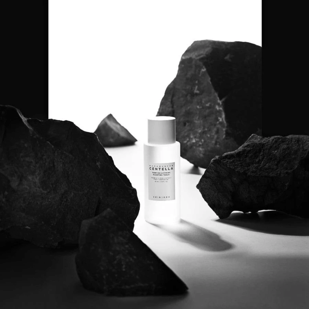 Weiße Flasche SKIN1004 Madagascar Centella Tone Brightening Boosting Toner  dramatisch platziert zwischen großen schwarzen Steinen vor weißem Hintergrund.