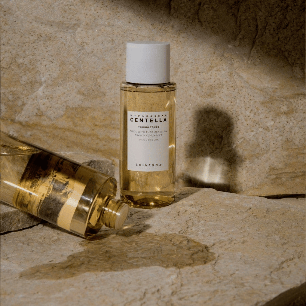 Das Bild zeigt eine Flasche "Madagascar Centella Toning Toner" von SKIN1004. Es ist eine transparente Flasche, gefüllt mit einer gelblichen Flüssigkeit, wahrscheinlich ein Toner für die Hautpflege. Es steht, dass sie mit reiner Centella aus Madagaskar hergestellt ist und der Inhalt beträgt 210 ml / 7,10 fl.oz. Die Flasche und eine liegende Flasche sind auf einer steinigen Oberfläche platziert, was ein natürliches und minimalistisches Gefühl vermittelt.