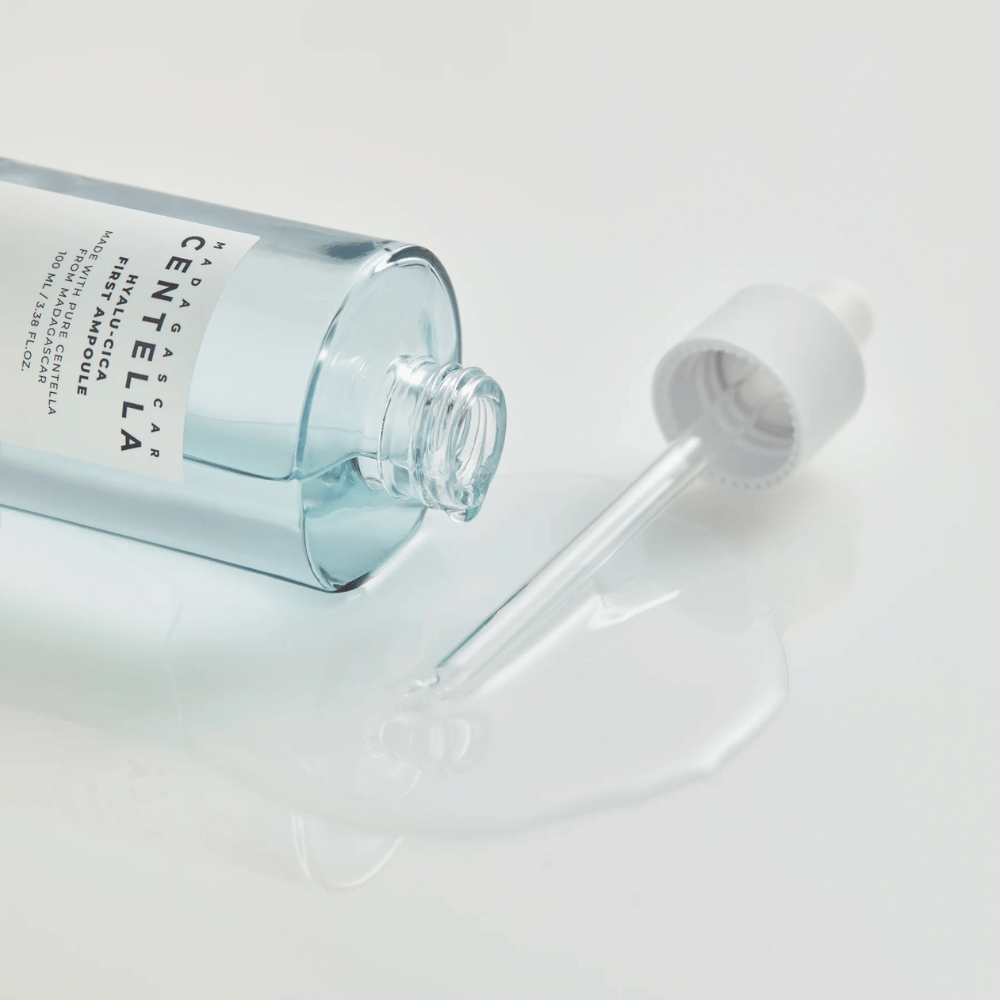 Nahaufnahme einer offenen Flasche Skin1004 Madagascar Centella Hyalu-Cica First Ampoule mit liegender Pipette und Serum, das auf einer weißen Fläche verteilt ist.