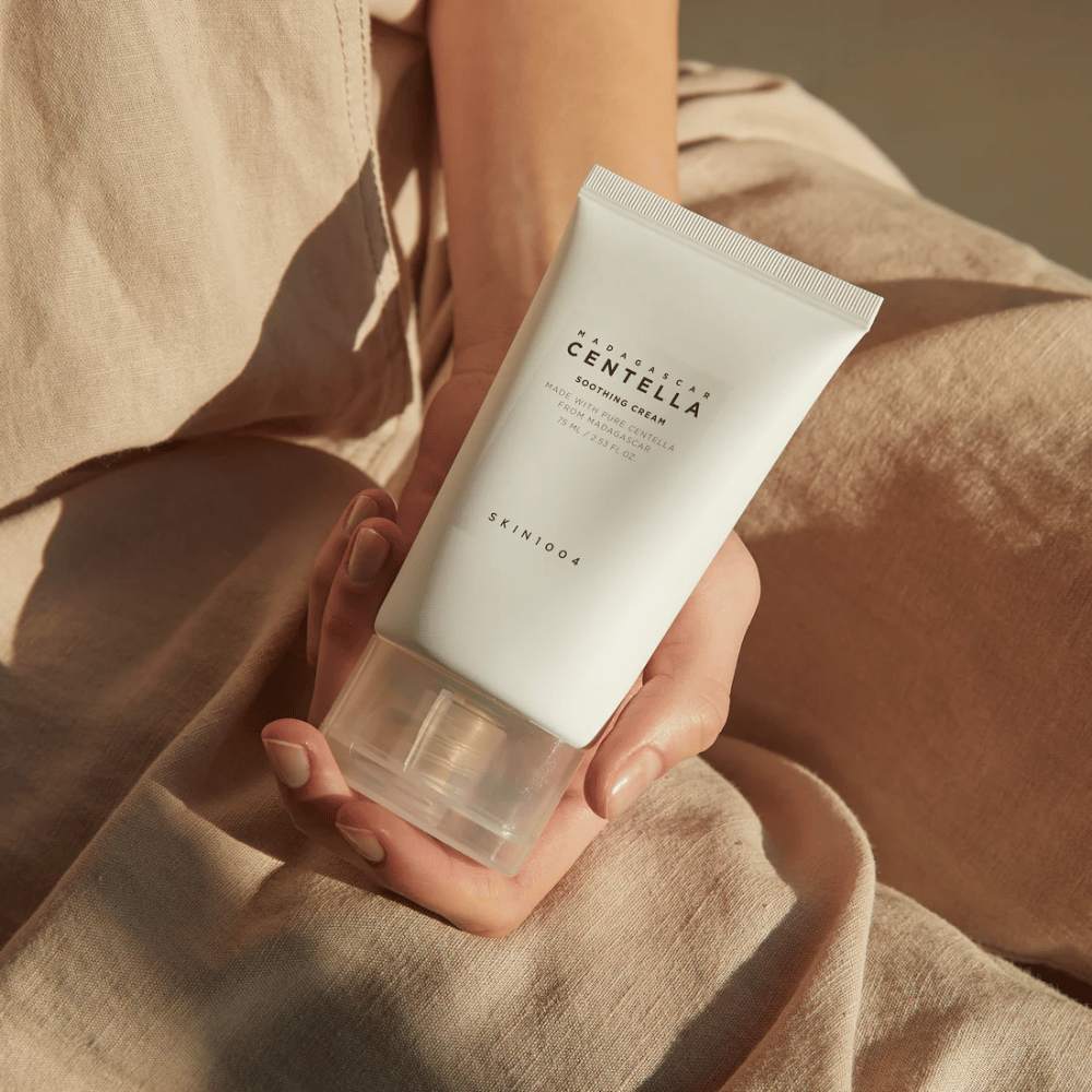 Eine Person hält eine weiße Tube von Madagascar Centella Soothing Cream aus SKIN1004. Die Tube hat einen durchsichtigen Verschluss und Text, der das Produkt als beruhigend und hergestellt mit reiner Centella beschreibt. Die Person trägt helle Kleidung.