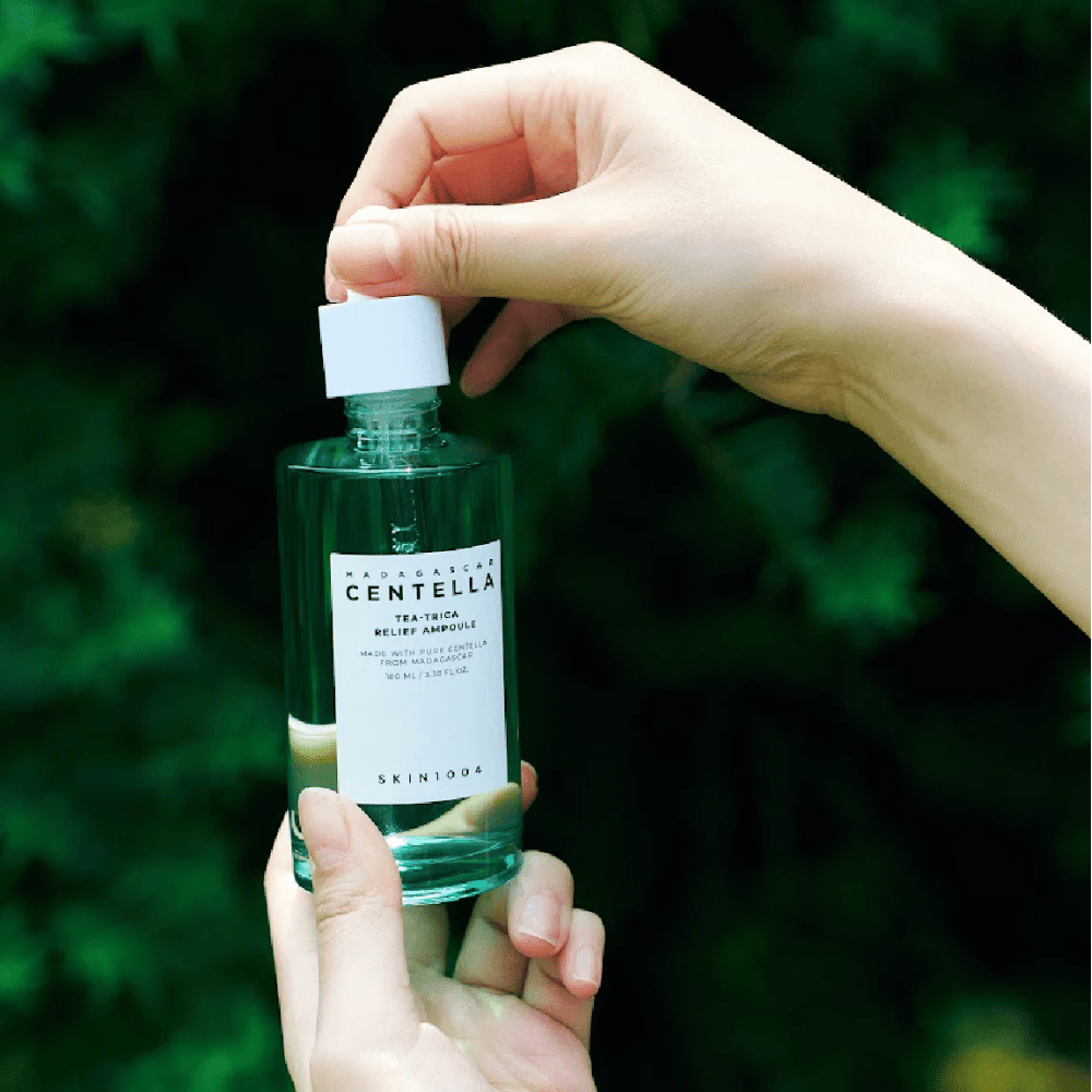 Person öffnet SKIN1004 Madagascar Centella Tea-Trica Relief Ampoule – grüne Glasflasche mit weißem Etikett wird in den Händen vor grünem Hintergrund gehalten.
