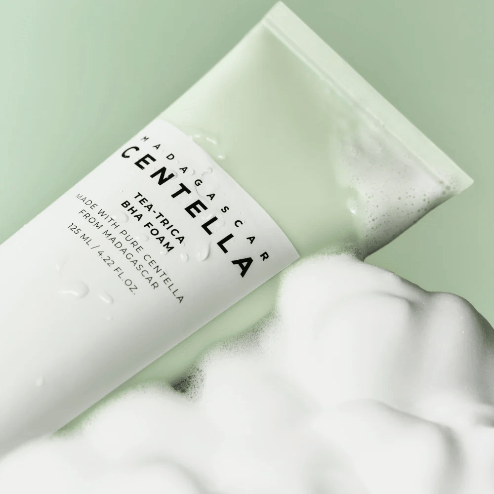 Ein Produkt vom Typ Reinigungsschaum, "Madagascar Centella Tea-trica BHA Foam", ist teilweise sichtbar mit Schaum drumherum. Das Produkt hat ein grünes und weißes Design, und der Text "Madagascar Centella" und "Tea-trica BHA Foam" sind deutlich sichtbar. Das Volumen beträgt 125 ml.