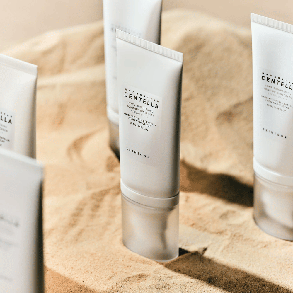 Mehrere Tuben mit Madagascar Centella Tone Brightening Tone-Up Sunscreen stehen im Sand. Die Tube ist weiß mit schwarzem Text und enthält 50 ml. Das Produkt hat SPF 50+ PA++++ und wird mit reiner Centella aus Madagaskar hergestellt.