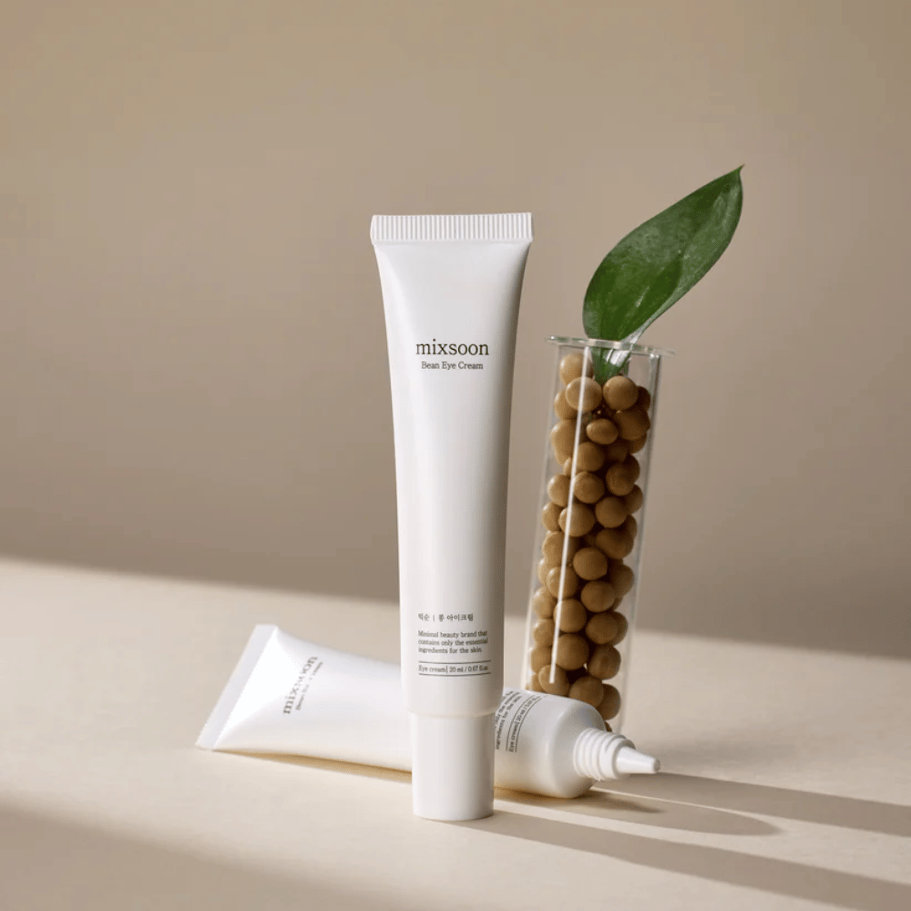 Zwei Tuben Mixsoon Bean Eye Cream neben einem Reagenzglas, das mit Sojabohnen gefüllt ist, und einem grünen Pflanzenblatt, auf einer beigen Oberfläche mit Schattenspiel.