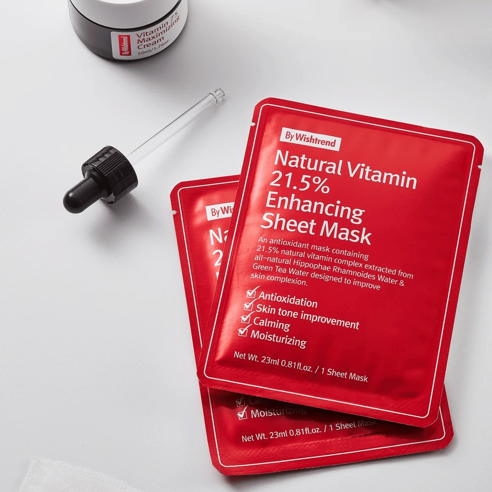 By Wishtrend Natural Vitamin 21.5% Enhancing Sheet mask in roten Verpackungen, platziert auf einer weißen Oberfläche zusammen mit einer Pipette und einem Glas Gesichtscreme.