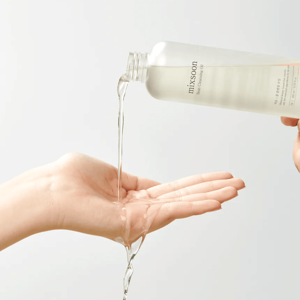 Ein klares Reinigungsöl wird aus einer transparenten Flasche mit weißem Etikett, auf dem "MixSoon" steht, in eine offene Handfläche gegossen. Das Öl bildet einen glänzenden Strahl zwischen der Flaschenöffnung und der Hand und demonstriert die flüssige Konsistenz des Öls. Die Hand ist ruhig und wird so gehalten, dass sie die gesamte Menge des Reinigungsöls vor einem einfarbigen, hellen Hintergrund auffangen kann.