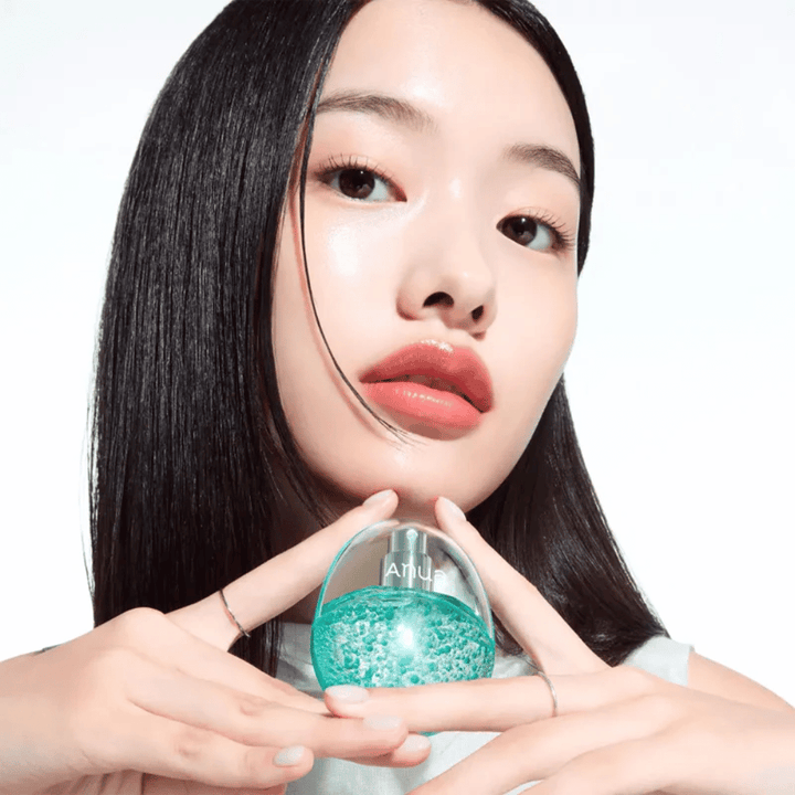 Nahaufnahme eines Models, das eine runde, transparente Anua-Flasche hält, die mit türkisfarbenen Kapseln oder Gelperlen gefüllt ist. Das Bild fokussiert den Glow der Haut und das innovative Design des Produkts, was einen frischen und modernen Eindruck vermittelt.
