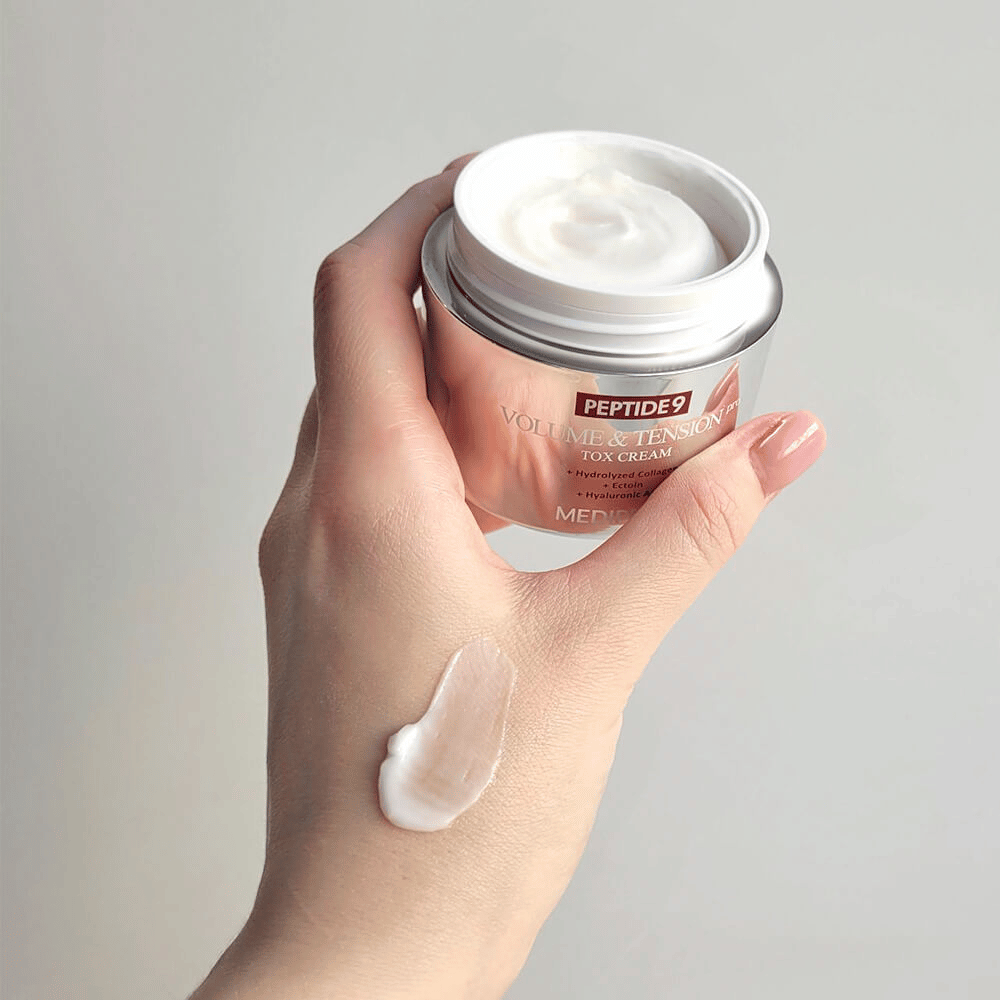 Hand hält die MEDI-PEEL Peptide 9 Volume & Tension Tox Cream mit einem Klecks Creme auf dem Handrücken. Die seidig weiche Formel spendet Feuchtigkeit und verleiht der Haut Festigkeit und Spannkraft.