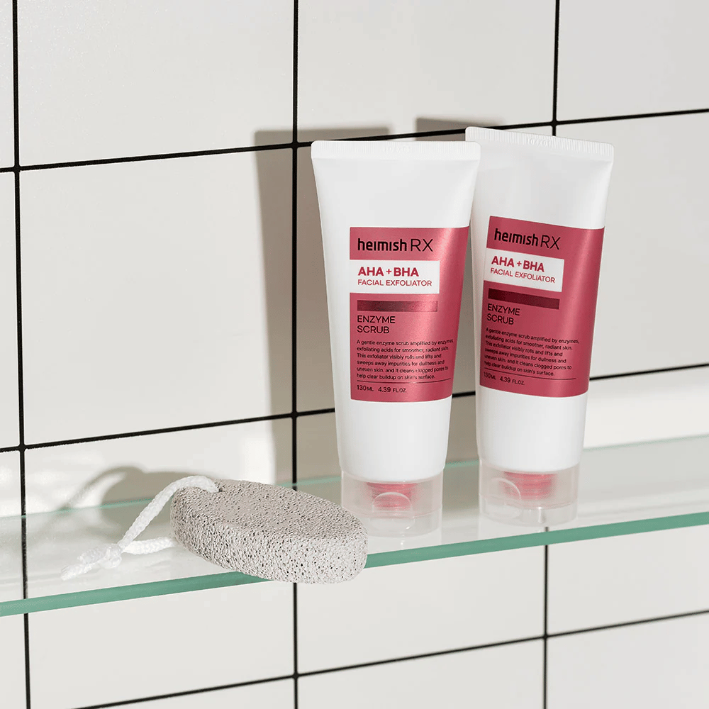 Zwei Tuben Heimish RX AHA + BHA Gesichtspeeling auf einem Glasregal in einem Badezimmer mit weißer Fliesenwand. Neben den Produkten liegt ein Bimsstein mit einem weißen Band. Ein stilvolles und minimalistisches Hautpflegebild.