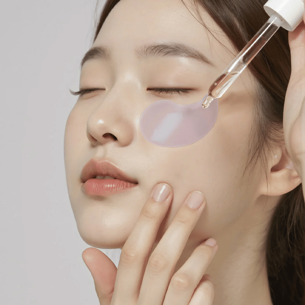 Eine Frau mit einem lila Hydrogel-Augenpad trägt Serum unter dem Auge mit einer Pipette auf. Das Produkt spendet Feuchtigkeit und pflegt die empfindliche Haut um die Augen.
