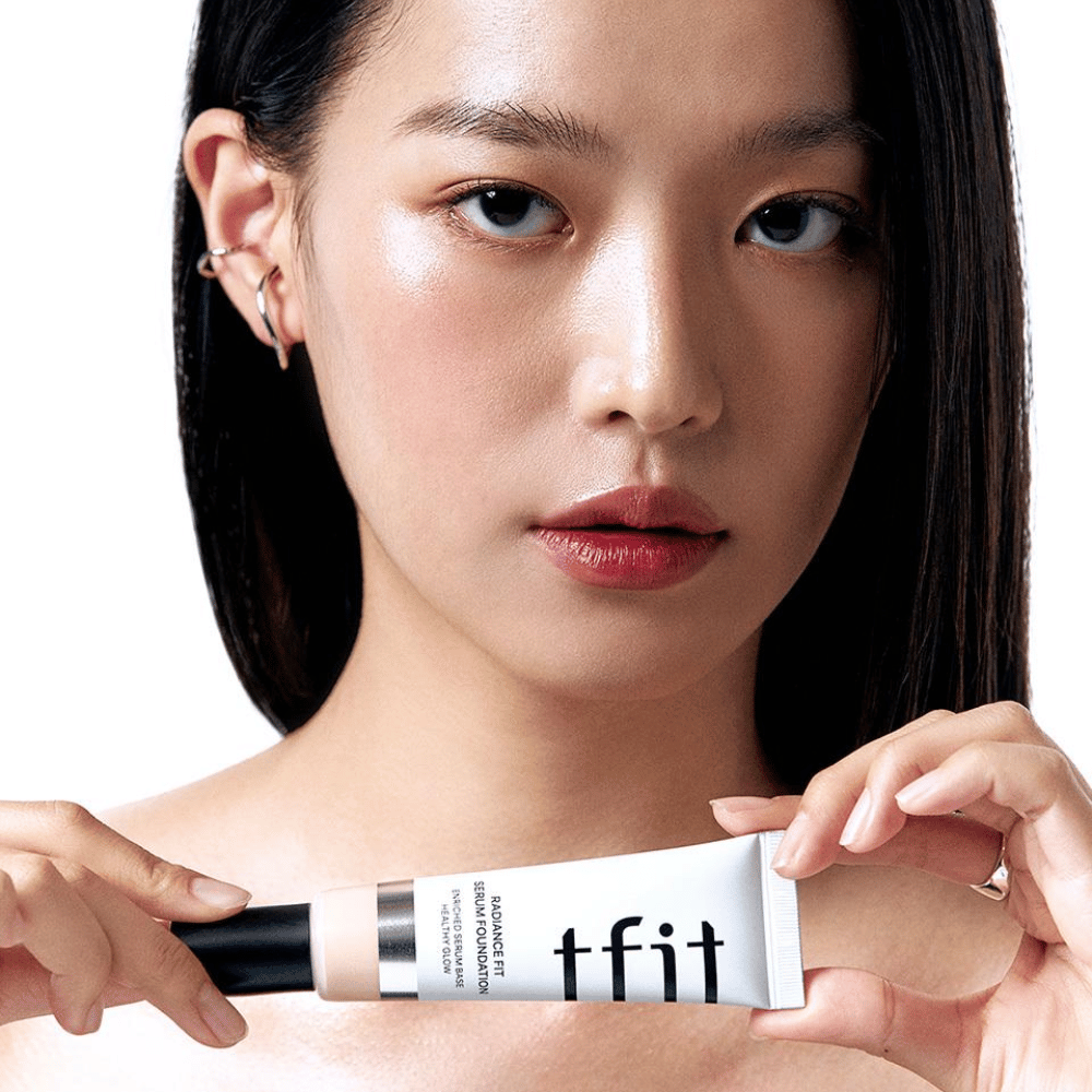 Modell mit glänzender, glatter Haut hält TFIT Radiance Fit Serum Foundation, was das natürliche Finish und den Glanz des Produkts hervorhebt.
