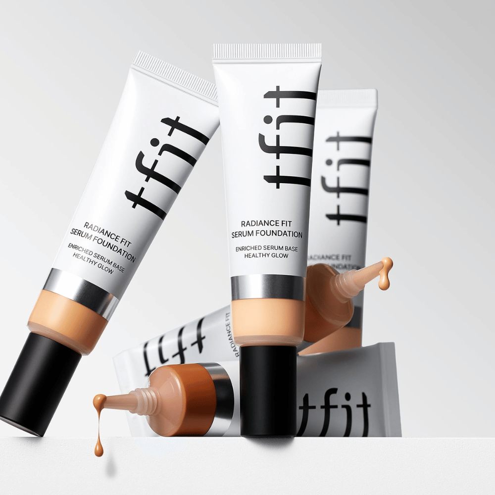Mehrere Tuben von TFIT Radiance Fit Serum Foundation in verschiedenen Nuancen, eine davon mit foundation, die heraus tropft, vor einem hellgrauen Hintergrund.
