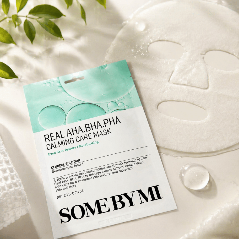 SOME BY MI Real AHA.BHA.PHA Calming Care Mask wird zusammen mit der entfalteten Tuchmaske gezeigt. Um das Produkt herum sind Feuchtigkeitstropfen und grüne Blätter zu sehen, die das Gefühl von Feuchtigkeitsversorgung, beruhigender Pflege und sanftem Peeling verstärken. Das Bild hebt den Fokus der Maske auf ein ebenmäßigeres Hautbild und eine verbesserte Feuchtigkeitsbalance hervor.