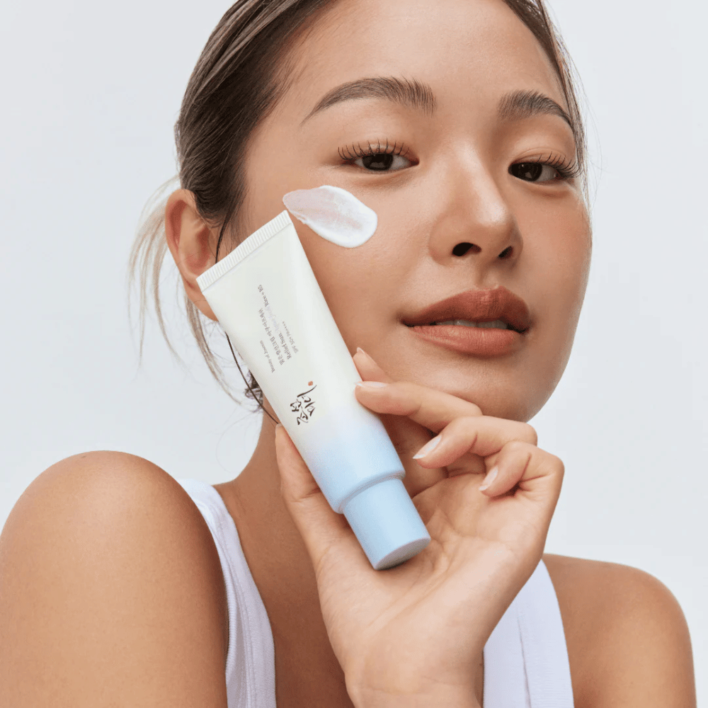 Frau hält Beauty of Joseon Relief Sun: Aqua-Fresh Rice + B5 Sonnencreme und zeigt deren cremige Konsistenz auf der Wange. Ein leichtes Sonnenschutzprodukt, das die Haut mit SPF 50+ PA++++ befeuchtet und schützt.