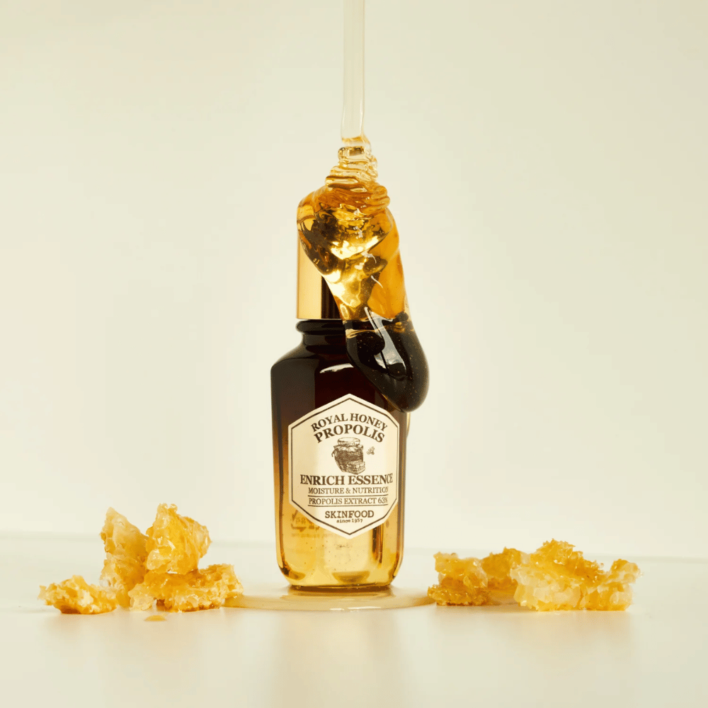 Bild von einer Glasflasche mit Royal Honey Propolis Enrich Essence von Skinfood. Ein großer Tropfen goldenen Honigs läuft vom goldfarbenen Deckel der Flasche herunter. Um die Flasche herum liegen kleine Stücke von Honigwaben auf einer hellen Oberfläche verstreut. Die Essenz wird mit einem Fokus auf Feuchtigkeit und Ernährung vermarktet, mit 63% Propolisextrakt.
