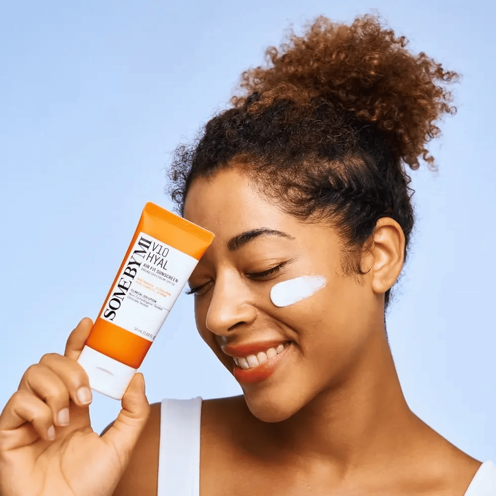 Eine Frau hält eine Tube mit SOME BY MI V10 Hyal Air Fit Sunscreen und hat einen weißen Fleck Sonnencreme auf der Wange. Sie lächelt fröhlich und zeigt das Produkt deutlich, was seine Anwendung und Vorteile für Hautpflege und Sonnenschutz hervorhebt.