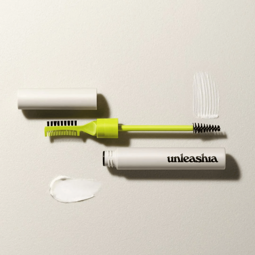 Ein Augenbrauenskulptur-Set mit einem grünen Kamm, einer schwarzen Bürste und einer weißen Tube mit der Aufschrift "unleashia" vor einem hellen Hintergrund mit einem Klumpen durchsichtiger Produkt.