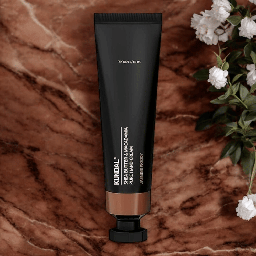 KUNDAL Shea Butter & Macadamia Pure Hand Cream Jasmine Woody auf Marmor mit Blumen platziert – nährende Handcreme, die trockene Hände pflegt und einen eleganten Jasmine Woody-Duft verleiht.