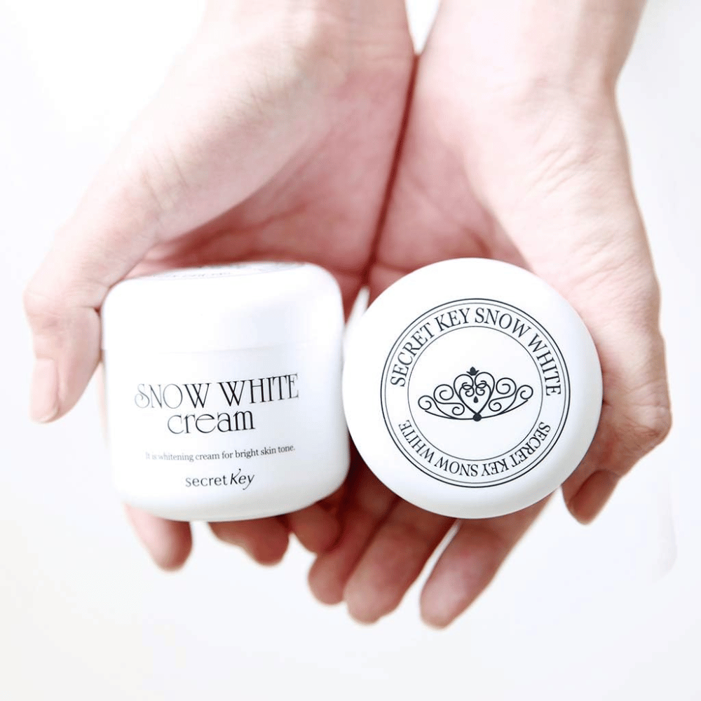 Zwei Dosen Secret Key Snow White Cream werden in den Händen gehalten, eine zeigt den Deckel und eine zeigt die Vorderseite.