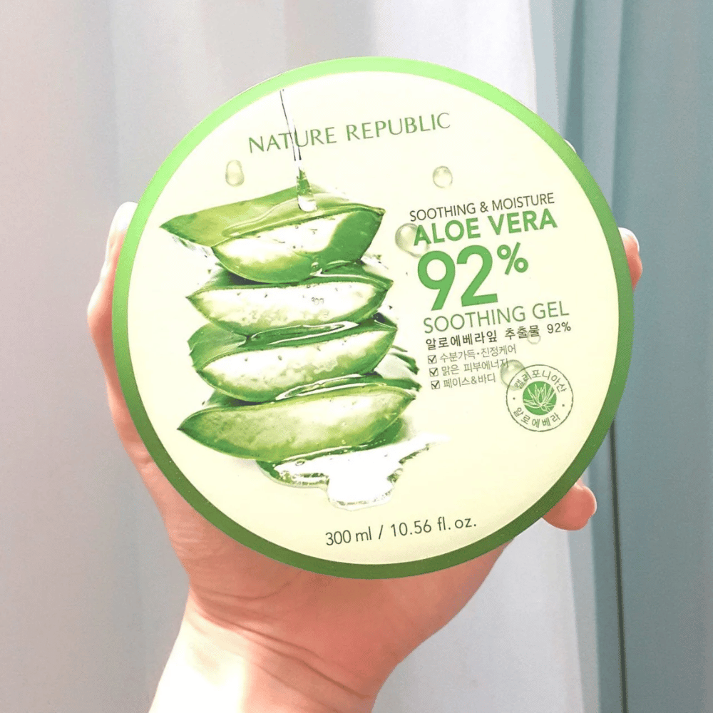 Eine Hand hält eine runde Dose mit Nature Republic Aloe Vera 92% Beruhigungsgel hoch. Die Dose ist grün mit einem Bild von Aloe Vera und Text, der ihre beruhigenden und feuchtigkeitsspendenden Eigenschaften beschreibt. Der Inhalt beträgt 300 ml.