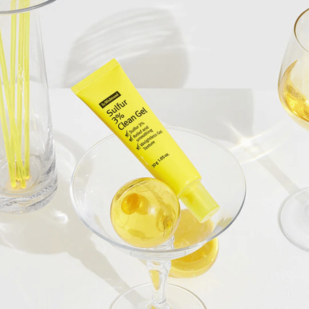 By Wishtrend Sulfur 3% Clean Gel in einer gelben Tube, platziert in einem Cocktailglas mit einer gelben Sphäre, umgeben von eleganten Gläsern auf einem weißen Hintergrund.