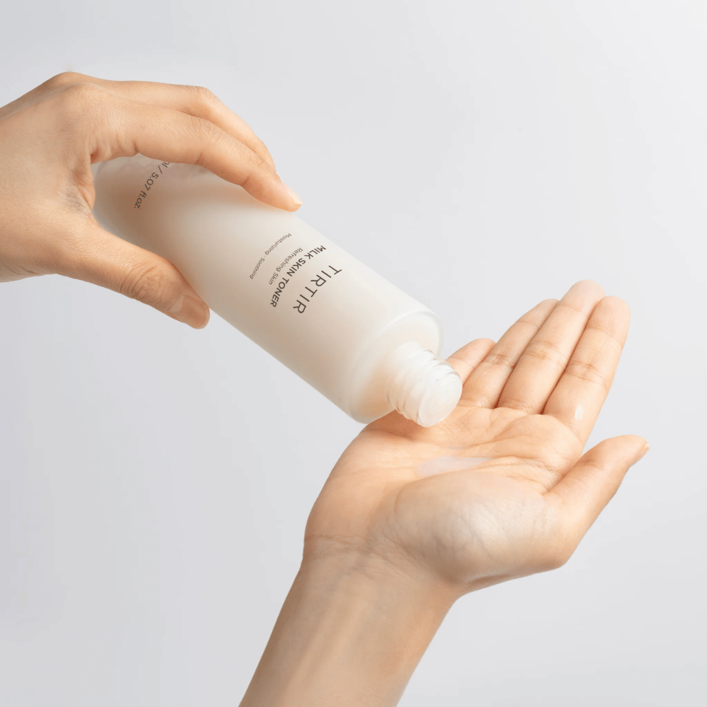 Das Bild zeigt eine Person, die eine milchige Flüssigkeit aus einer Flasche mit der Aufschrift "TIRTIR Milk Skin" auf die Handfläche gießt. Das Produkt scheint ein Hauttoner oder eine feuchtigkeitsspendende Lotion zu sein.