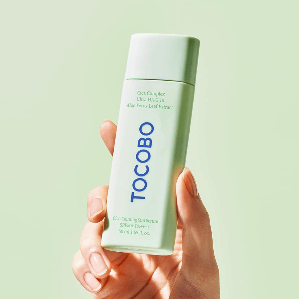 Hand hält TOCOBO Cica Calming Sun Serum mit mintgrüner Verpackung und blauer Schrift vor hellgrünem Hintergrund.