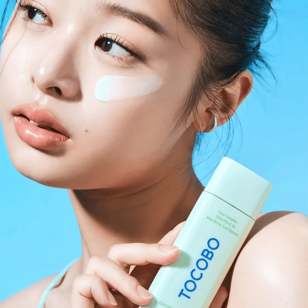 Modell mit Sonnencreme unter dem Auge hält TOCOBO Cica Calming Sun Serum in mintgrüner Verpackung vor blauem Hintergrund.