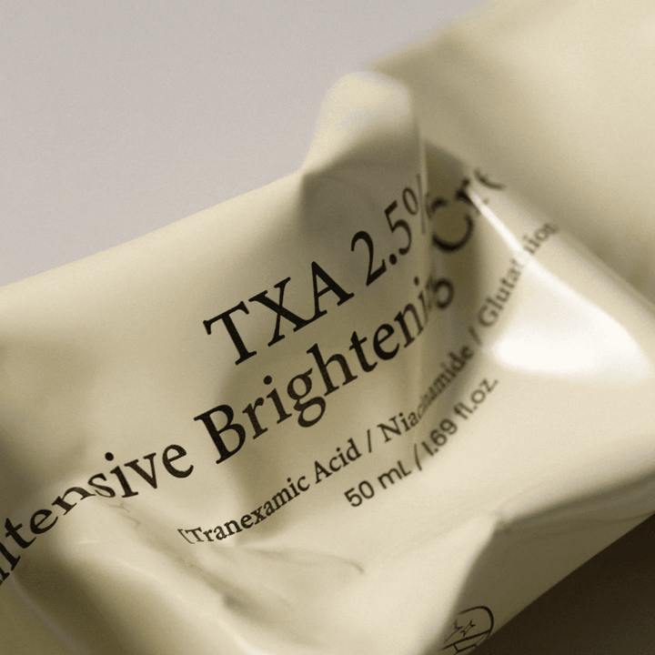 Nahaufnahme von AXIS - Y TXA 2.5% Intensive Brightening Cream, bei der das weiche und minimalistische Tubendesign im Fokus steht. Das Bild hebt den Produktnamen, Tranexamic Acid und Niacinamide sowie die elegante beige Verpackung hervor.