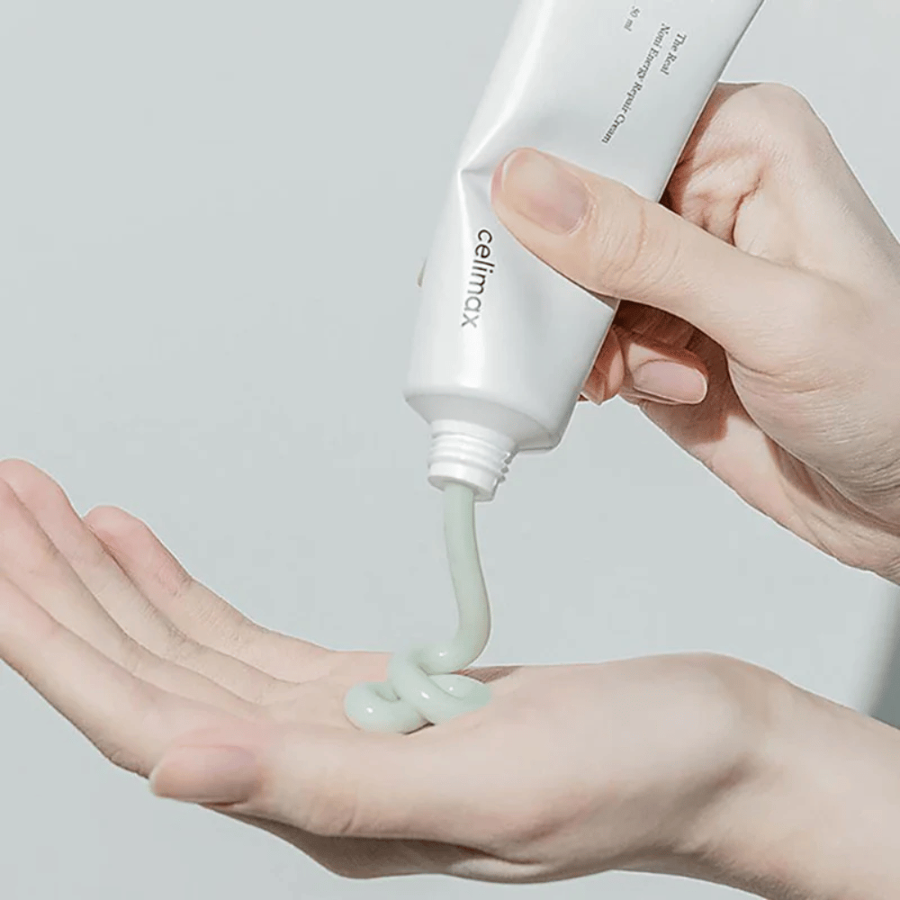 Eine Hand hält eine weiße Tube mit Celimax Noni Energy Repair Cream, und die Creme wird in einem gleichmäßigen Strahl auf die Handfläche gedrückt. Die Creme ist hellgrün und wird zur Pflege und Wiederherstellung der Haut vermarktet.
