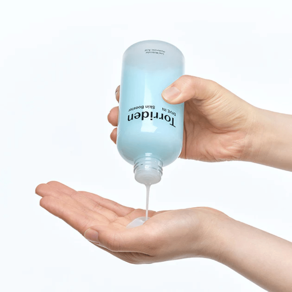 Hände, die eine Flasche mit Hautpflegeprodukt halten und eine klare Flüssigkeit in die Handfläche dosieren. Die Flasche ist etikettiert mit "Torriden - DIVE-IN Low Molecule Hyaluronic Acid Skin Booster".