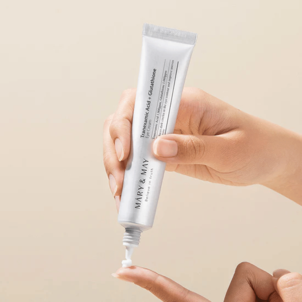 Eine Hand hält eine Tube MARY & MAY "Tranexamic Acid + Glutathione Eye Cream". Die Tube ist weiß und das Design ist sauber und einfach, was die beruhigenden und aufhellenden Eigenschaften des Produkts für den Augenbereich hervorhebt. Das Produkt wird sanft aufgetragen, mit Fokus auf Präzision und Effizienz.