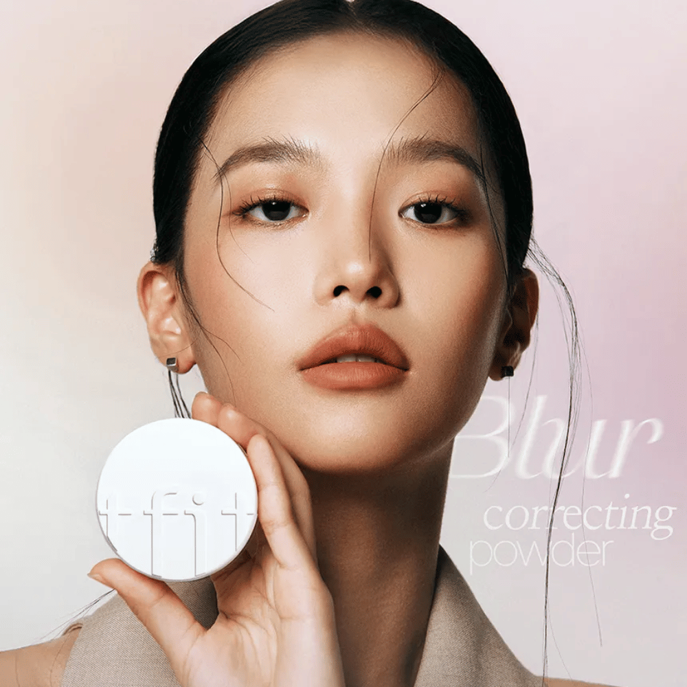 Frau mit makellosem Make-up hält eine runde weiße Dose mit TFIT-Logo hoch, mit dem Text "Blur Correcting Powder" im Hintergrund.