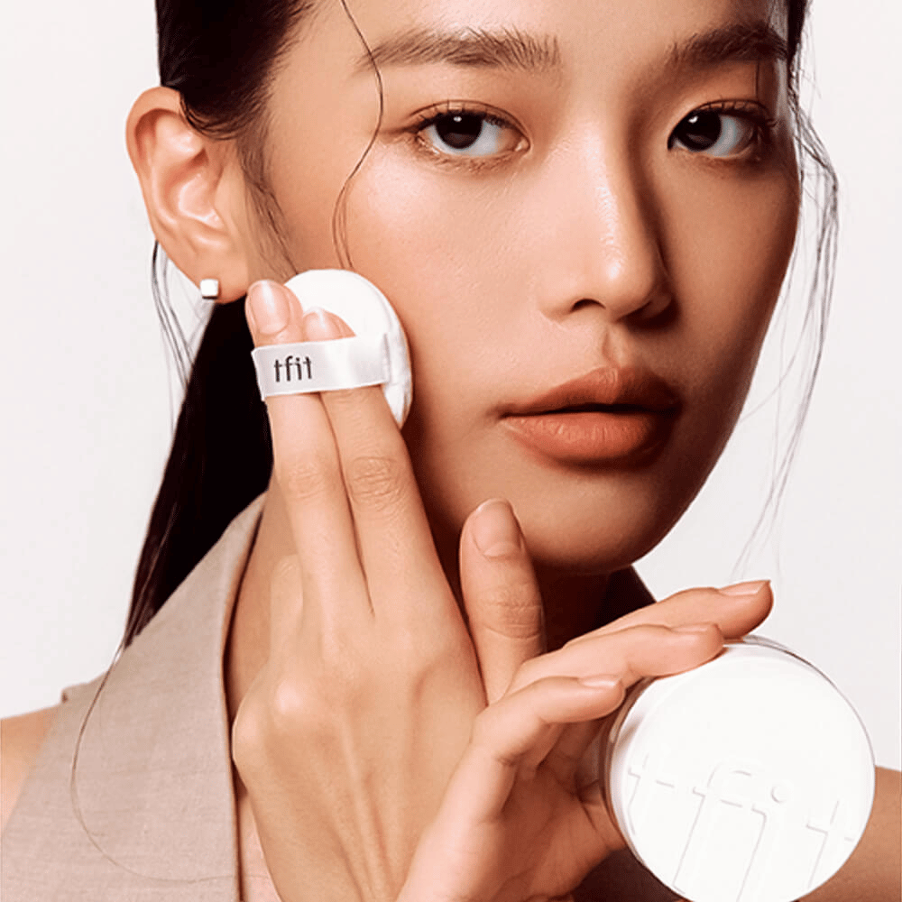 Frau trägt TFIT Translucent Set Finishing Powder #02 Skin Beige mit einem Puff auf, hält eine weiße Puderdose mit TFIT-Logo.