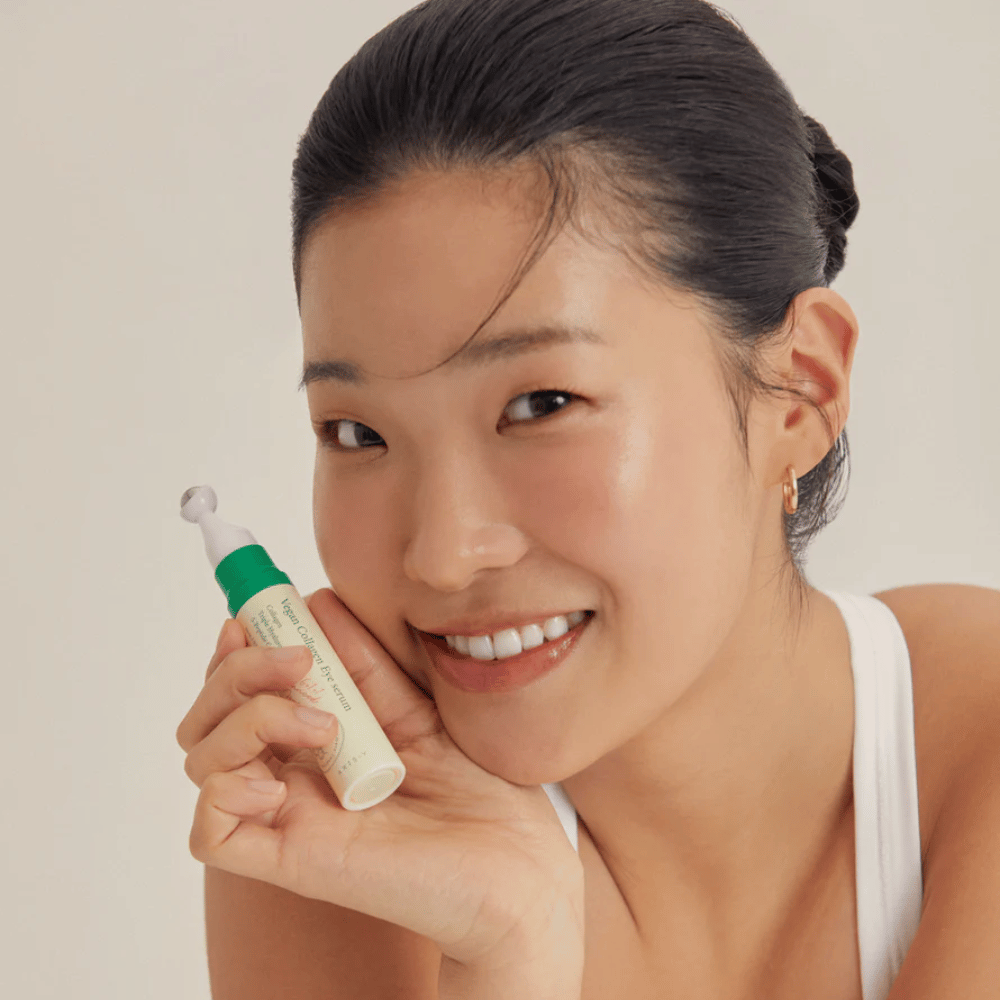 Eine Frau mit hochgestecktem Haar und goldenen Ohrringen hält ein AXIS-Y Vegan Collagen Eye Serum 10ml in der Nähe ihres Gesichts, während sie sanft lächelt. Das Serum ist entwickelt, um Feuchtigkeit zu spenden und Falten um die Augen zu reduzieren, speziell für empfindliche Haut entwickelt.