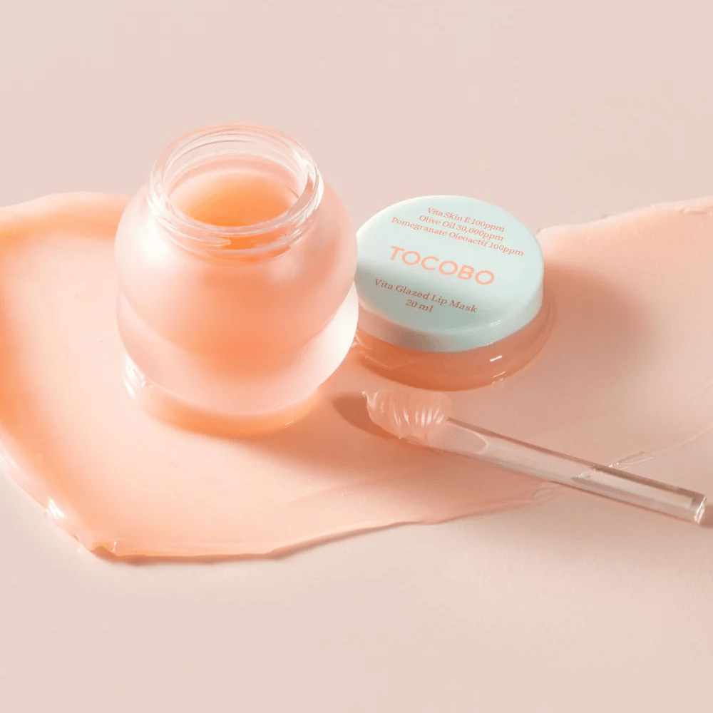 Ein Glas mit "Vita Glazed Lip Mask" von Tocobo wird angezeigt. Das Glas ist durchsichtig mit einem helltürkisen Deckel und enthält eine glänzende, rosa Lippenmaske. Ein kleiner Spatel liegt neben dem Glas, und ein Teil der Maske ist auf der Unterlage verteilt. Das Produkt ist dafür konzipiert, die Lippen mit Feuchtigkeit zu versorgen und zu pflegen.