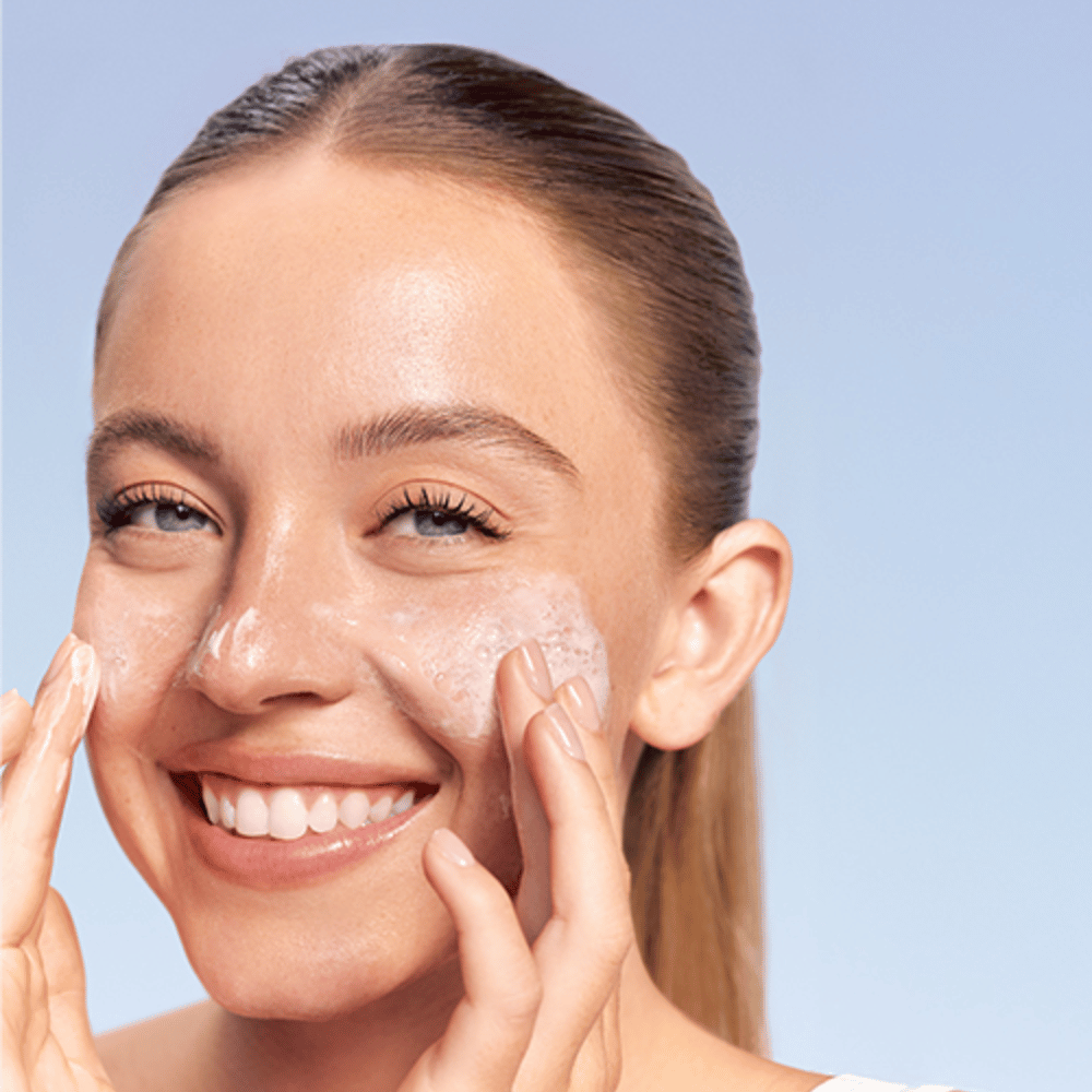 Lächelnde Frau trägt LANEIGE Water Bank Gentle Gel Cleanser in schäumender Form auf die Wangen auf, vor blauem Hintergrund.