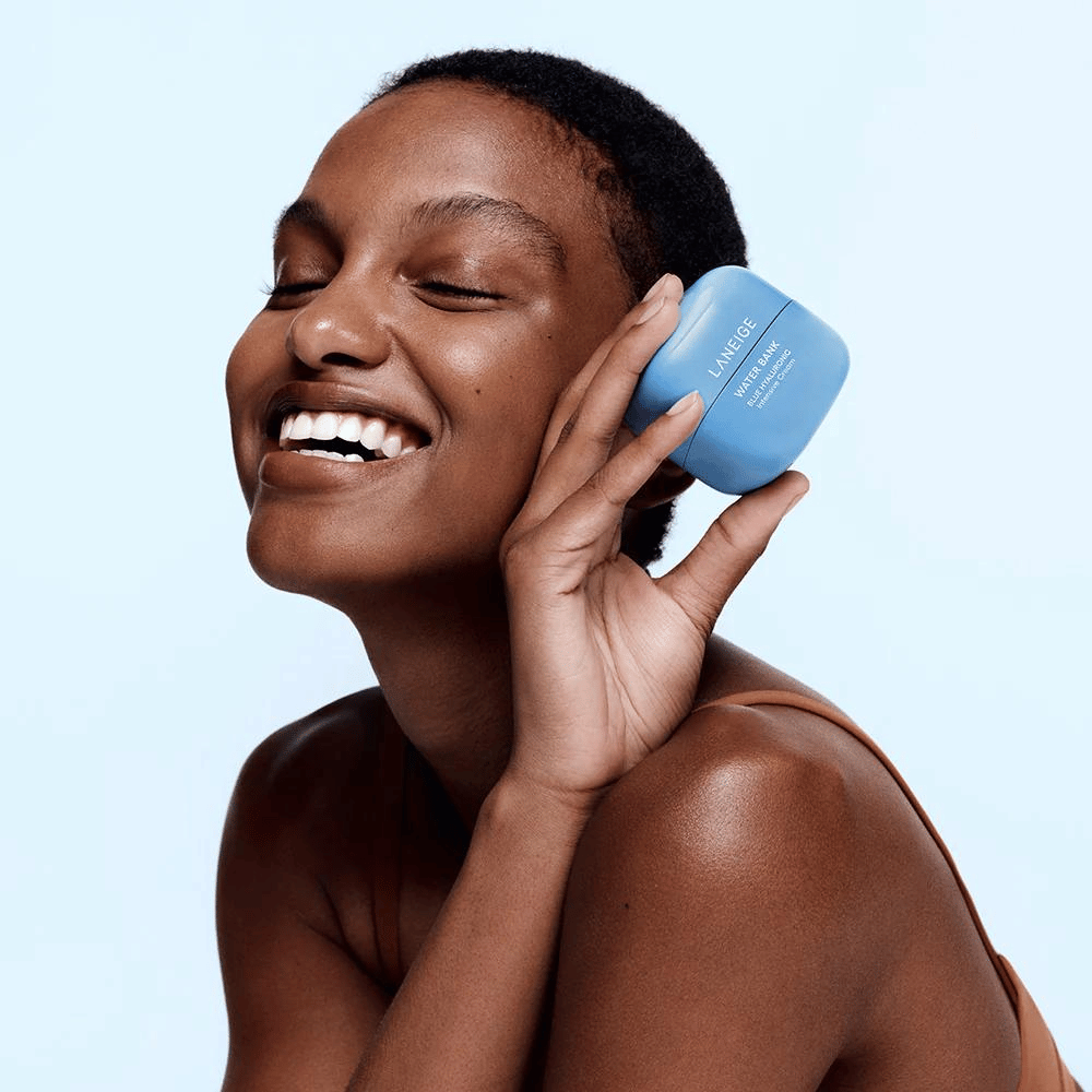 Lächelnde Frau hält eine blaue Dose LANEIGE Water Bank Blue Hyaluronic Cream an ihre Wange, vor hellblauem Hintergrund.