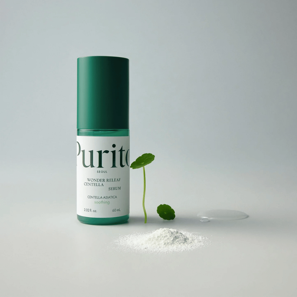Grüne Flasche von Purito Wonder Releaf Centella Serum 60 ml steht auf einem weißen Untergrund, umgeben von Centella-Blättern, einem durchsichtigen Serumtropfen und einem kleinen weißen Pulver.