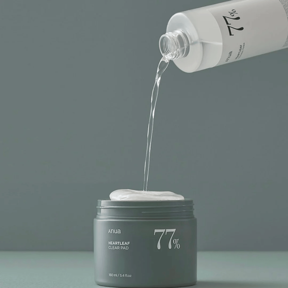 Eine durchsichtige Flasche gießt eine Flüssigkeit in einen grauen Behälter mit der Aufschrift "anua HEARTLEAF 77% Clear Pad", der einen Stapel weißer Hautpflegepads enthält. Der Hintergrund ist schlicht und einfarbig in einem sanften Grauton.