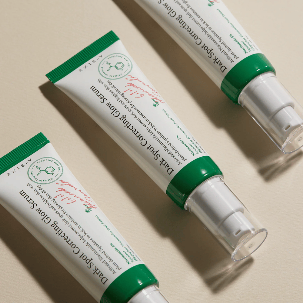Drei Tuben von Axis-Y Dark Spot Correcting Glow Serum mit grünem Verschluss und weißer Verpackung liegen auf einem hellen Untergrund.